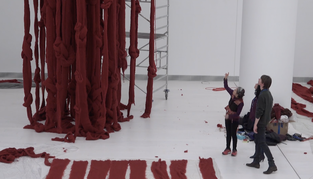 Aerial view into a white gallery where three people are installing and discussing a large red textile installation that hands from the ceiling