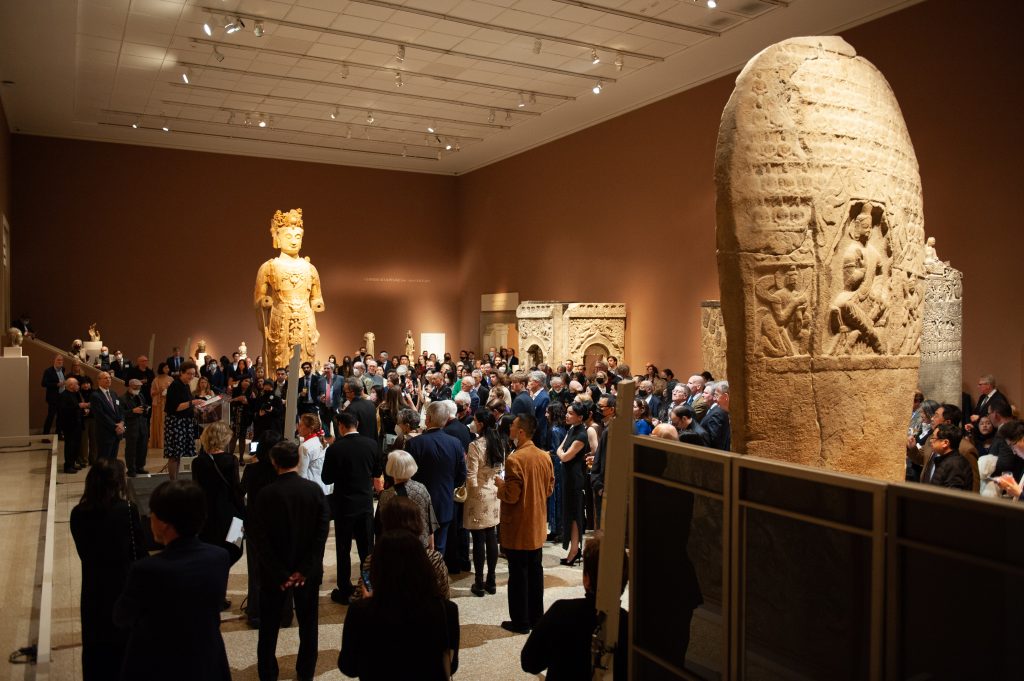 In a color photograph, finely attired people attend a party in a large gallery with brown walls and huge sculptures.