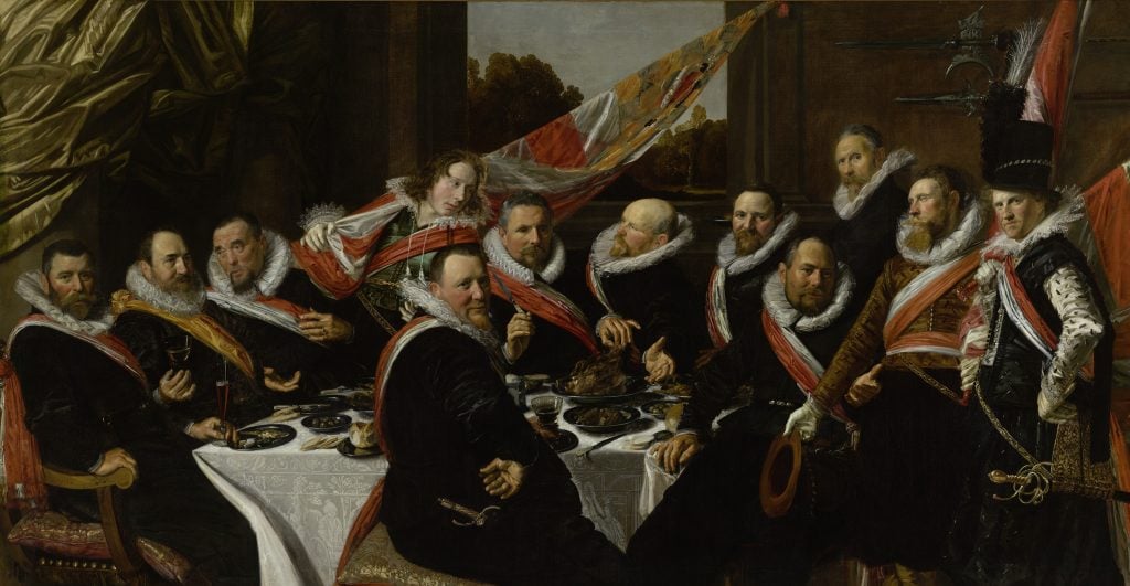 In a color photo of a painting, a large group of men in fancy dress sit around a table, enjoying fine food.