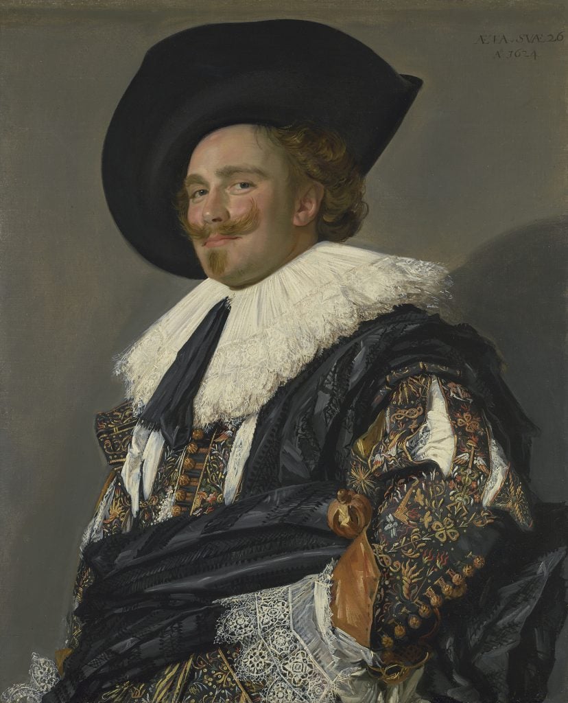 In a color photo of a painting, a man is seen from the waist up in an elaborate formal dress with fringed collar and sleeves and a black hat.