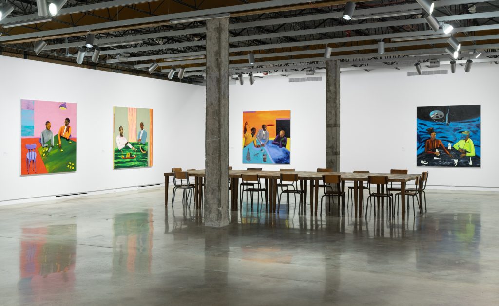 An art show by lubaina himid where there are several brightly colored paintings on the wall depicting figures and a large table with chairs in the center of the room