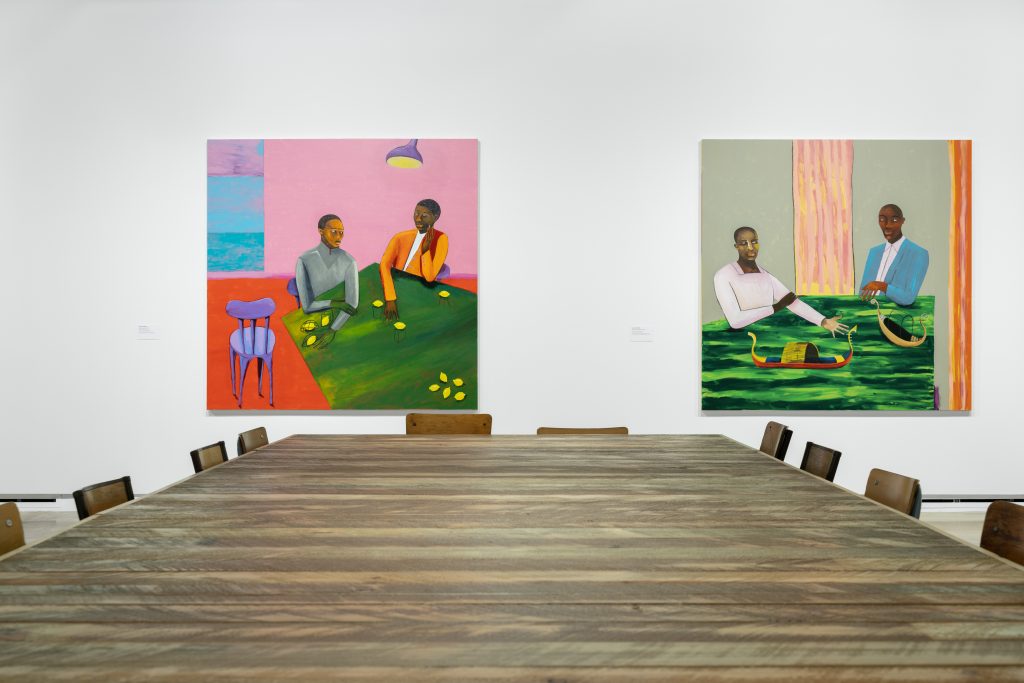 Art installation by Lubaina Himid that includes a large table and chairs and on the wall are two paintings in bright colors depicting figures seated at tables