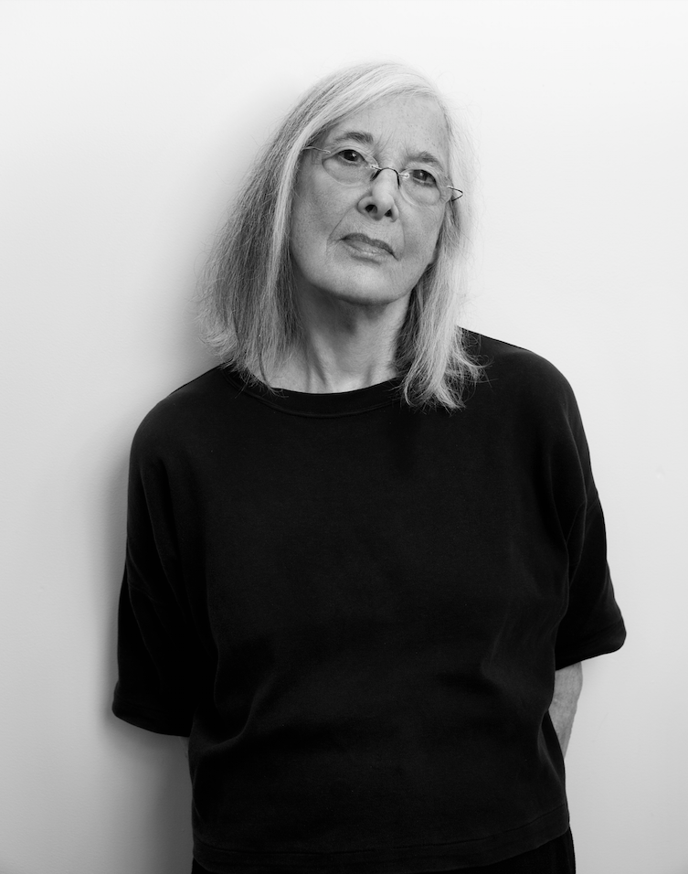 An older white woman with medium length gray hair looks out. She wears a dark. oversize shirt.