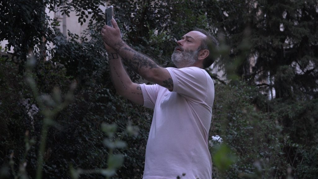 A bearded man in a white shirt taking a picture with his cellphone in the midst of greenery