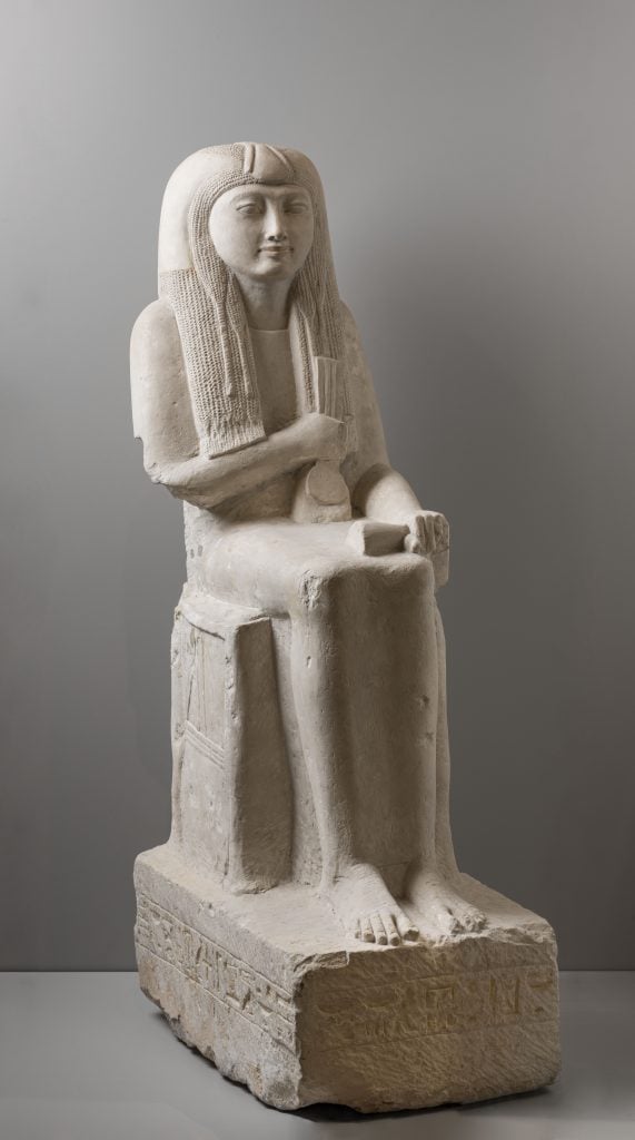 An ancient Egyptian statue of a woman sitting on a chair, holding ceremonial tools