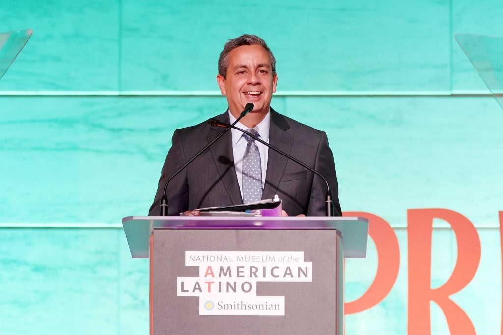 The image depicts a man standing at a podium and speaking. He is wearing a dark suit, a light-colored shirt, and a dotted tie. The podium features the logo of the National Museum of the American Latino, as well as the Smithsonian Institution logo. Behind him is a bright teal background, and there is some pink text visible to the right side that appears to be part of a larger word or phrase. The man is smiling and seems to be engaged in giving a speech or presentation. The setting suggests a formal event, likely related to the museum or a function hosted by the Smithsonian Institution celebrating or discussing aspects of Latino heritage in America.
