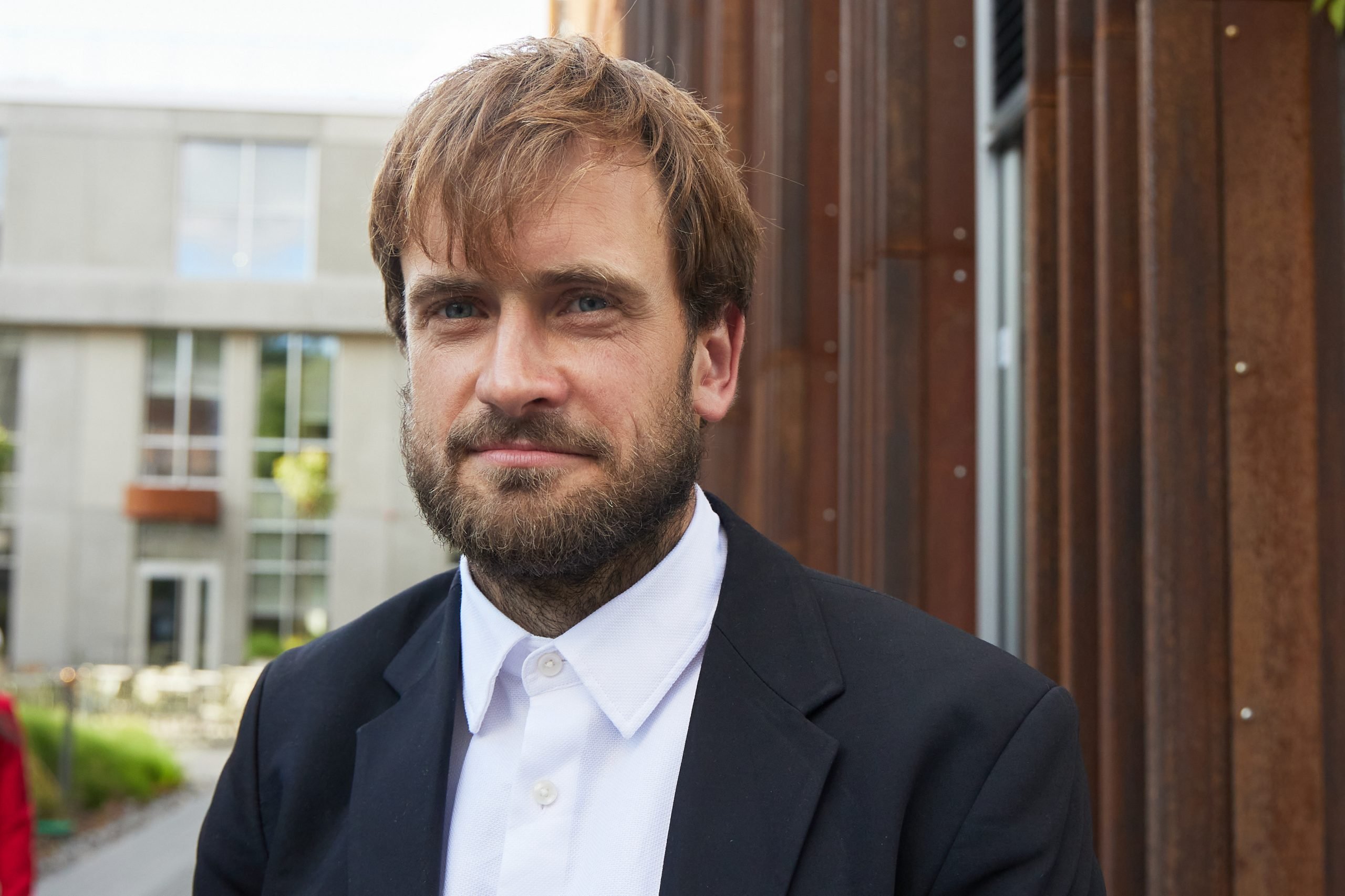 The image is a photo of a man in a suit, identified as the artist Pyotr Verzilov. He is wearing a black suit and is standing outdoors. The man has a beard and a mustache.