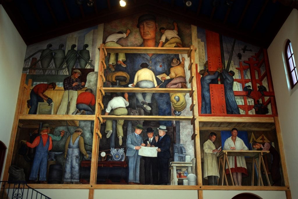This image is a photograph of a large mural painted on a wall. The mural is divided into several panels, each depicting different scenes of industrial and manual labor. The central figure in the top middle panel is a large, stoic male figure looking straight ahead, possibly representing the everyman of the labor force. The left side of the mural shows workers involved in carpentry and construction, with one man sawing a piece of wood, while others handle different types of equipment. The top-left corner has figures carrying what appear to be bags on their back, perhaps symbolizing the burden of labor. The right side of the mural has a prominent red structure, where workers are climbing and working on steel beams, reminiscent of the iconic imagery of skyscraper construction in the early 20th century, particularly in the United States. In the bottom middle panel, there are three men in suits and hats examining a document, representing the management or planning aspect of industrial work. To the right of this, there is a panel with two figures in lab coats, suggesting scientific or medical professions, with one figure writing notes and the other conducting an experiment or examination. The mural's style is characteristic of social realism, a genre that emphasizes realistic depictions of everyday life and often highlights themes of social justice and labor. The use of muted earth tones and the portrayal of the workers in a dignified, yet stark manner are typical of this style. It seems to convey a sense of the importance of the working class and the collective effort involved in industry and progress. The mural is housed within a room with a wooden ceiling and a balcony with a railing visible in the lower part of the image. The setting appears to be a public or institutional building, possibly a museum, hall, or even a former industrial building converted for public use.