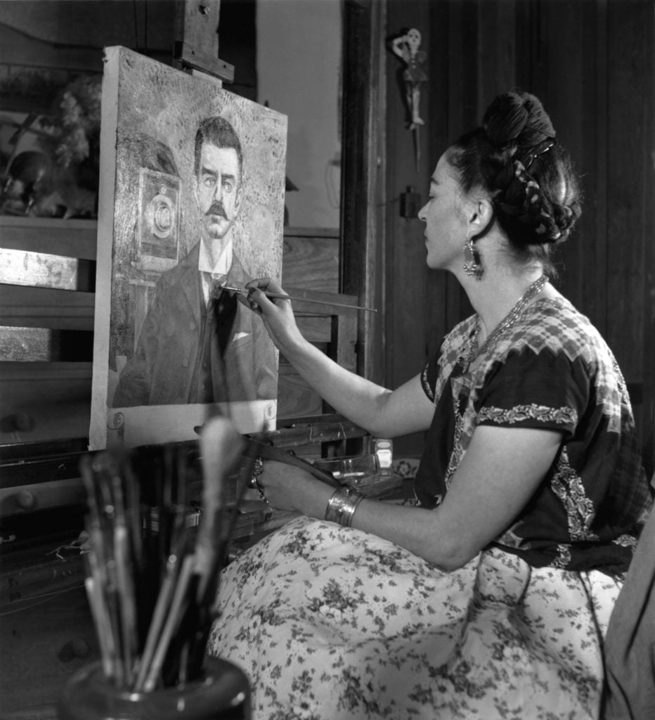 Mexican painter Frida Kahlo in a Tehuana dress painting a portrait of a mustachioed man