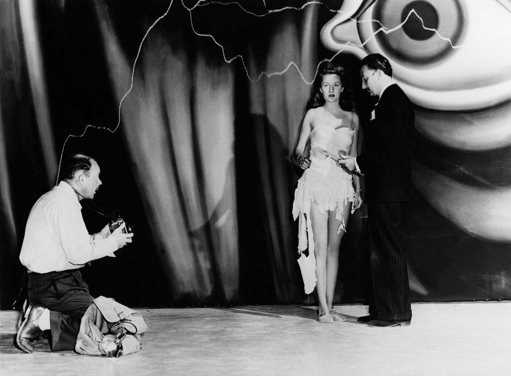 A black and white photograph of a man filming a woman in a white dress and a man in a black suit on a film set with a large eyeball in the background