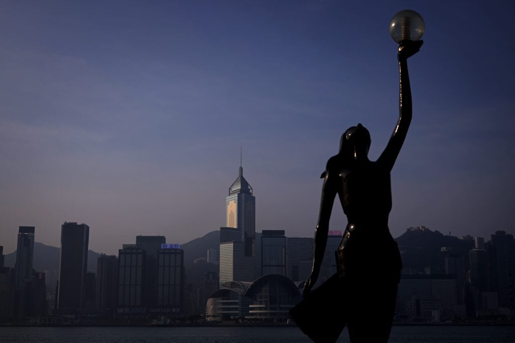 The image shows a statue of a woman holding a lamp in Hong Kong. The setting appears to be outdoors in the city.