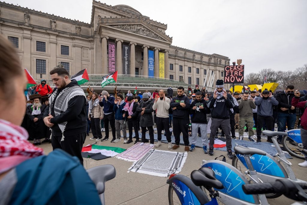 The image shows a group of people standing in front of the Brooklyn Museum at a protest. The individuals are wearing various clothing and are walking along a street in a crowd, waving Palestine flags. The setting is outdoors with a clear sky visible in the background.