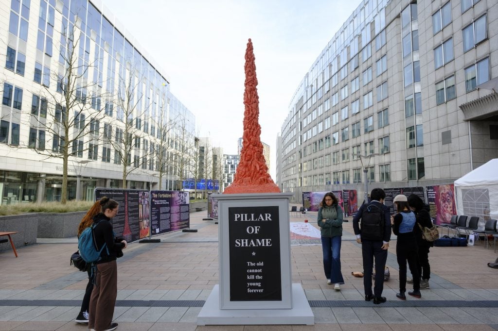 The image shows an orange statue in a plaza between buildings with many windows. The statue has a plaque with the text "Pillar of Shame: The old cannot kill the young forever." It is located in a town square with pedestrians in the background.