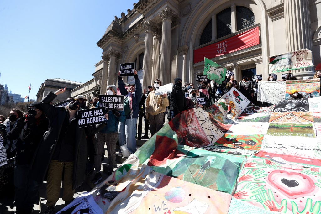 The image shows a group of people holding signs and flags in front of the Metropolitan Museum of Art in New York. The signs displayed express support for Palestine and freedom. The protestors are advocating for the right of return and a free Palestine.