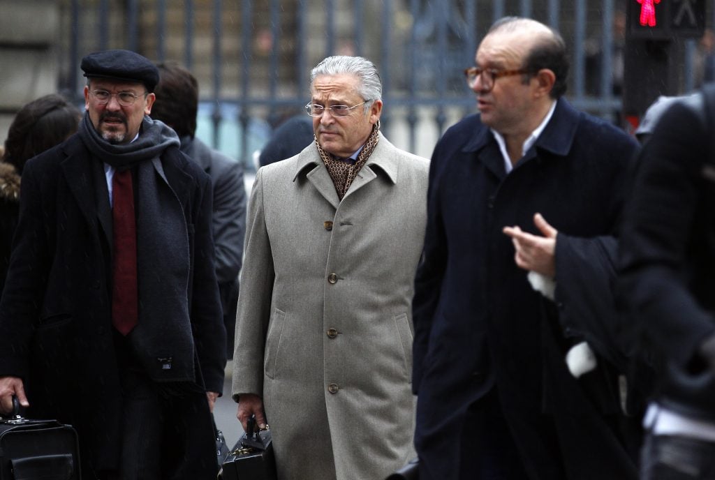 This photograph appears to show three men walking side by side. The man in the center is older, wearing a light beige coat over what seems to be a leopard print scarf and glasses. He carries a briefcase, has gray hair, and is wearing a pair of glasses. The man to his left (in the picture) wears a dark coat, a beret, a red tie, and also carries a briefcase. The man to the right is wearing a dark navy coat and has his hand raised, as if in mid-gesture, possibly in conversation with the other two men. They are all dressed formally, suggesting they could be on their way to a business meeting or legal proceeding, given the briefcases and their serious expressions.