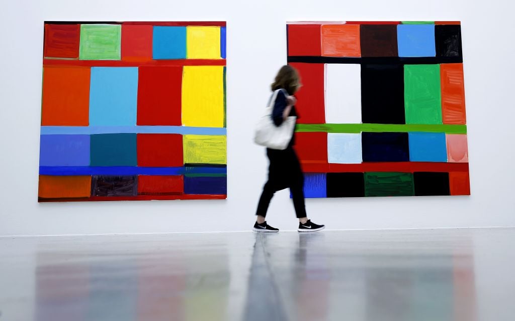 The image features a person walking by two large colorful grid paintings on a white wall.