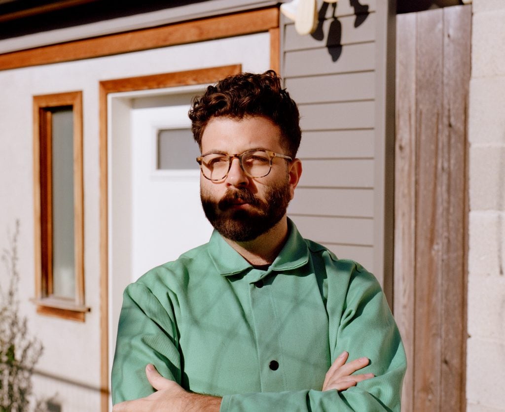 Artist Sam Ghantous standing in front of a doorway in a green shirt with his arms crossed looking into the distance.