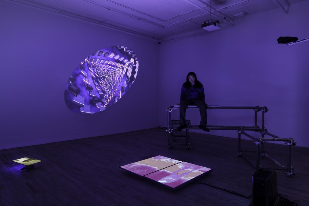 A gallery room filled with blue-purple light and a screen on the floor projecting an amorphic shape in space.