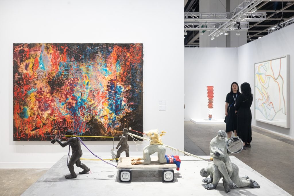 In a color photo a vibrant painting hangs on a white wall and a variety of childlike sculptures sit on a white plinth in the foreground.