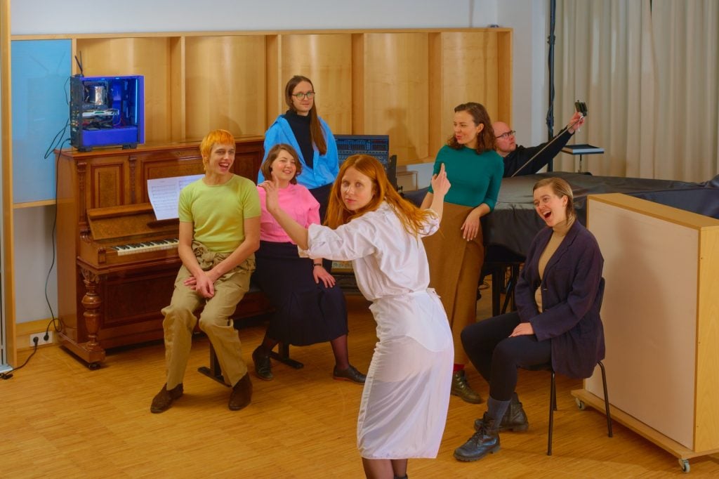 The image is of a group of people posing for a photo indoors. The people are smiling and wearing various types of clothing. There is a piano, desk, chair, and computer in the room where the photo is being taken.