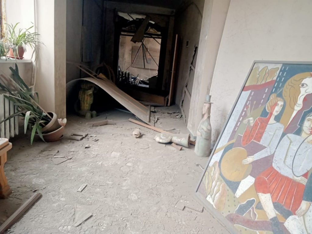 An art studio corridor covered in dust and debris with a warped metal structure, paintings, and a broken sculpture, indicating a scene of devastation