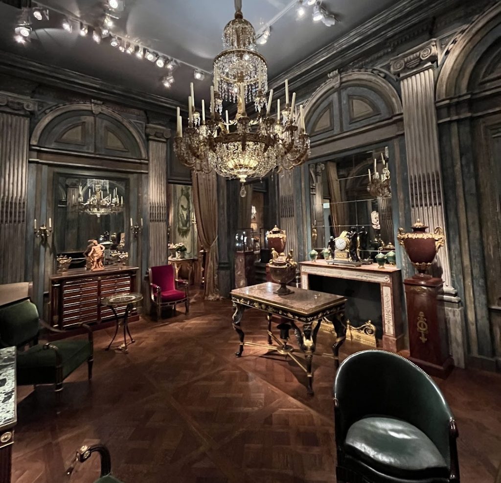 In a color photo, an opulent interior is seen with a chandelier, wood and leather furniture, and more.
