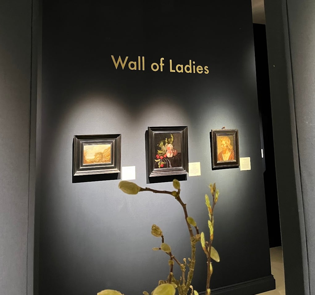 A color photo shows three small paintings on a gray wall. The phrase "Wall of Ladies" is written on the wall.