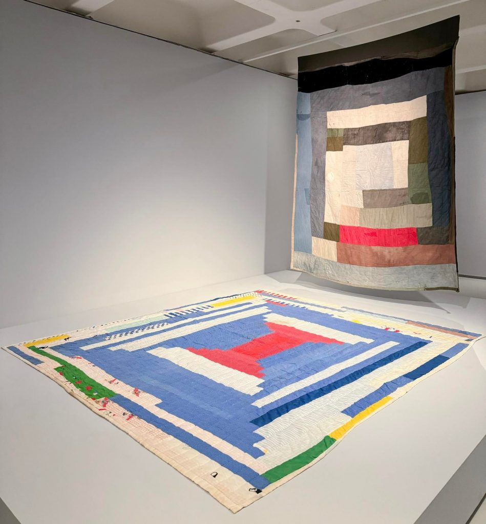 a quilt flat on a raised platform and another quilt hanging above it, abstract patterns on both
