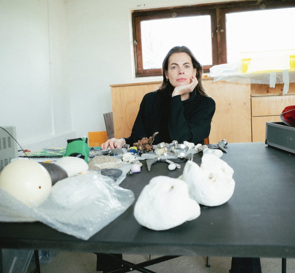 a person, the artist Saskia Noor van Imhoff, sitting at a table with a group of white objects on it. The person appears to be smiling. The setting is indoors against a wall.
