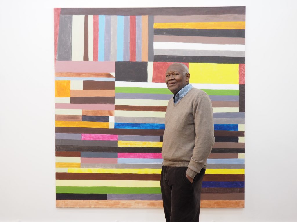 The image shows artist Atta Kwami standing in front of his painting, which features multiple colorful stripes placed horizontally and vertically. Kwami is wearing trousers and is standing against a wall.