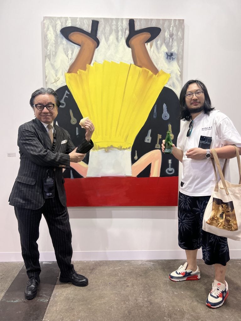 two me, one in a suit one very casual with long hair, pose with an artwork of a woman standing on her head in a yellow skirt. 