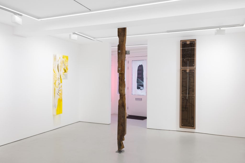 The image depicts a white room with a few abstract works on on the wall and a wood beam running floor to ceiling. The room appears to be an indoor art exhibition space with a white plaster floor and ceiling.