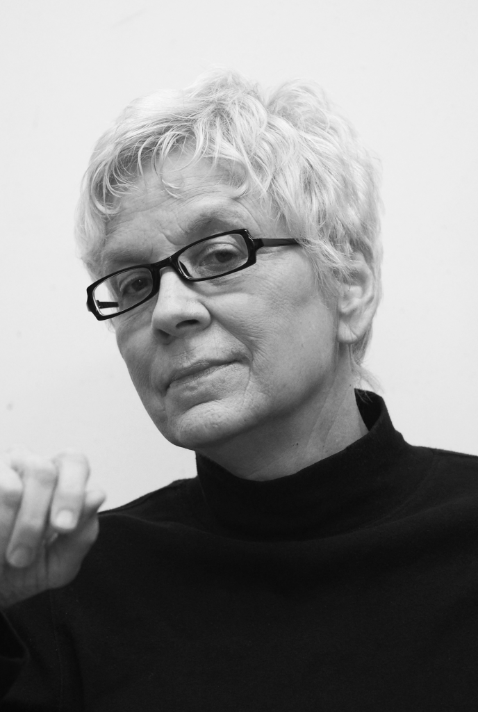 an older white woman with short white hair looks directly at the camera. she wears black framed glasses and a turtleneck.