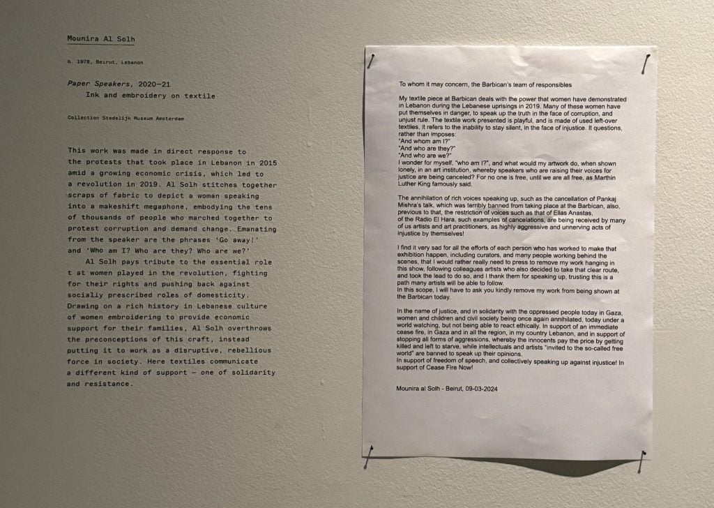 A piece of paper with typed writing is pinned next to text describing an artwork on a gallery wall.