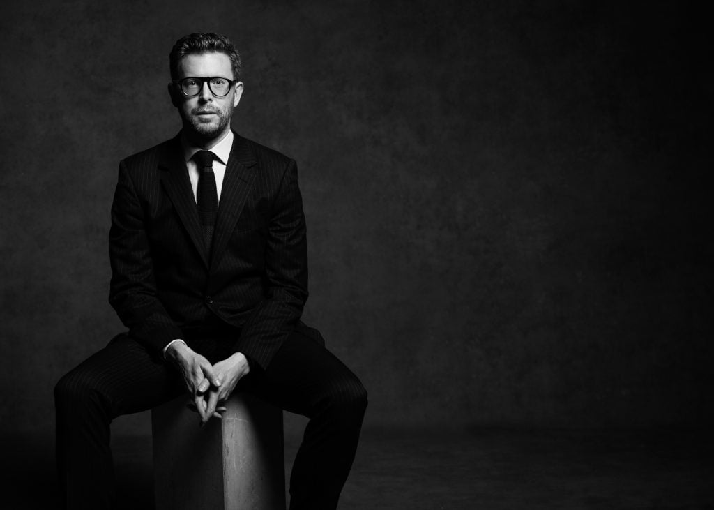 The image depicts a man sitting on a chair. He is wearing a blazer and glasses, and the photo is in black and white.