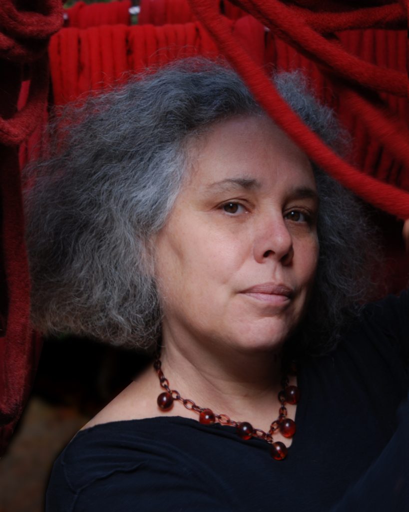 The image features a person with shoulder-length curly grey hair, gently framed by what appears to be a thick, red, rope-like structure, possibly part of a hammock or artistic installation. The person is looking directly at the camera, exuding a calm and thoughtful expression, and is wearing a black top with a scoop neckline. Accentuating the attire is a tasteful necklace with red beads, complementing the red structure around them. The background is somewhat indistinct