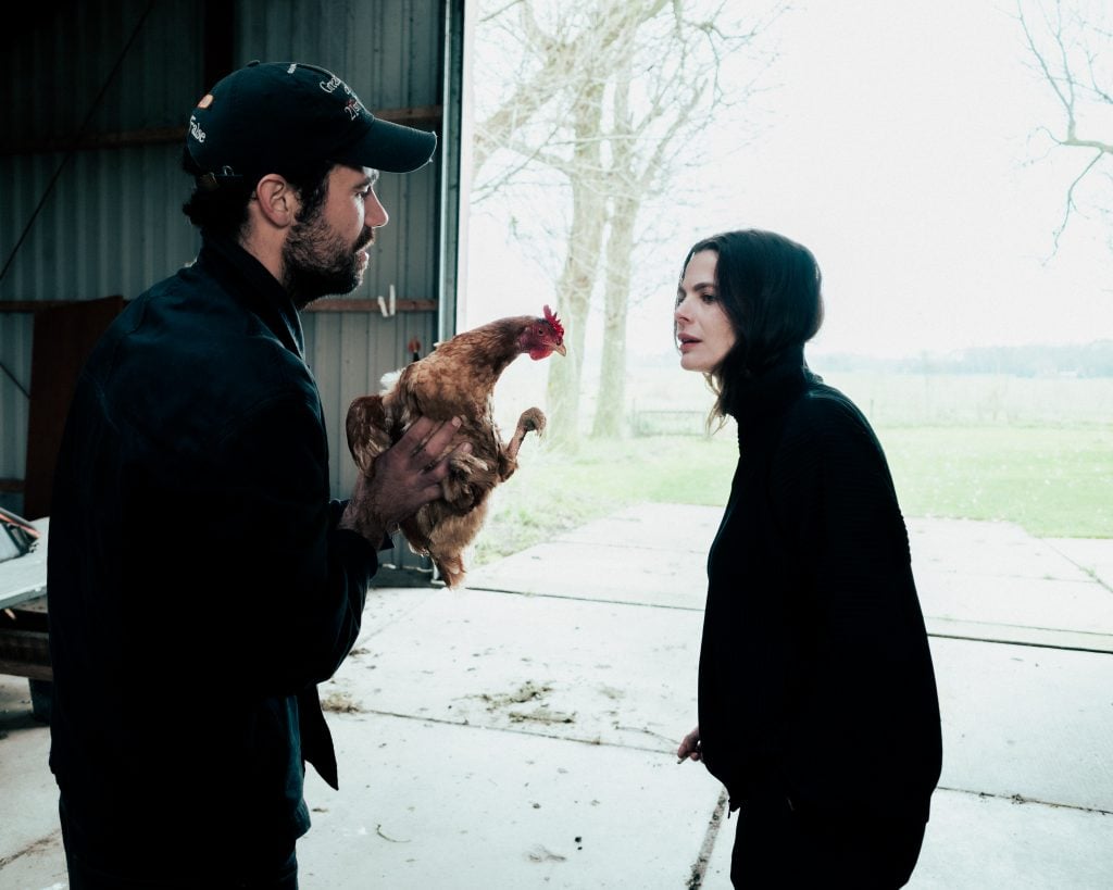 A man holds up a chicken while the artist Saskia Noor van Imhoff looks at the chicken. They are standing in a barn with the doors open.