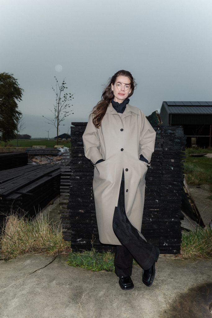 a woman, the artist Saskia Noor van Imhoff, wearing a trench coat outdoors, smiling, standing in front of a barn. The background includes a foggy sky, trees, and grass.