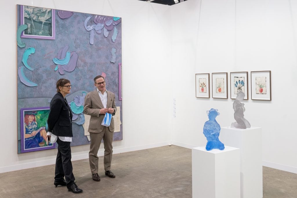In a color photo, a vibrant purple painting is on a white wall. Two people stand in front of it on a concrete-colored floor. A few other artworks are off to the side.