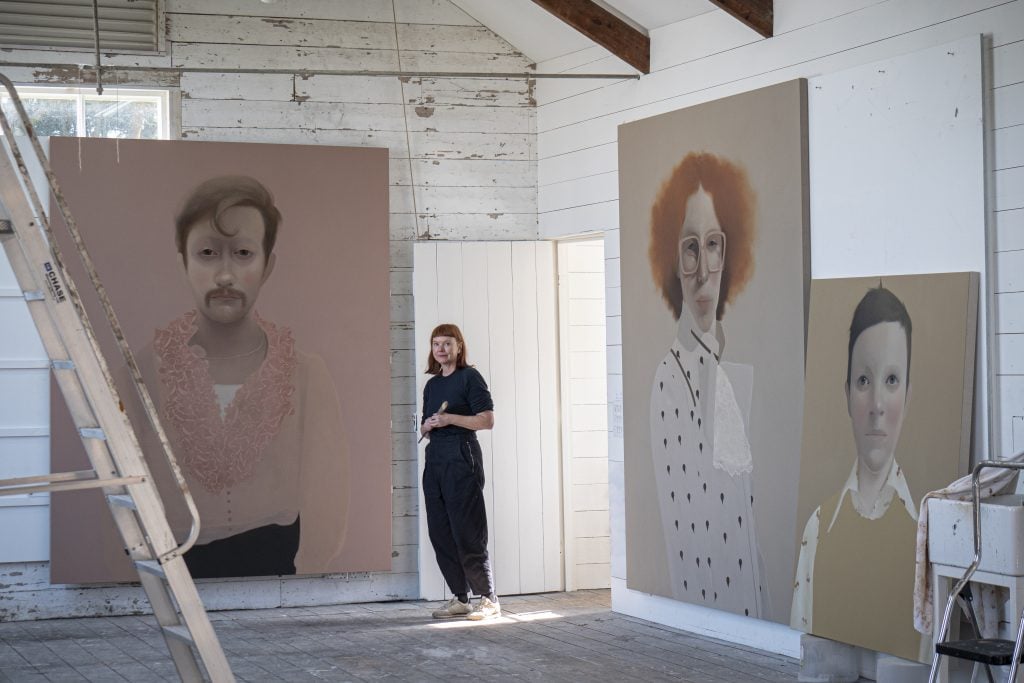 Artist Sarah Ball wearing black long-sleeve t-shirt, black pants, and sneakers in her studio surrounded by three large-scale paintings of individuals. Studio has a ladder in the foreground and is painted in distressed white, with the ends of vaulted ceiling beams showing at the top.