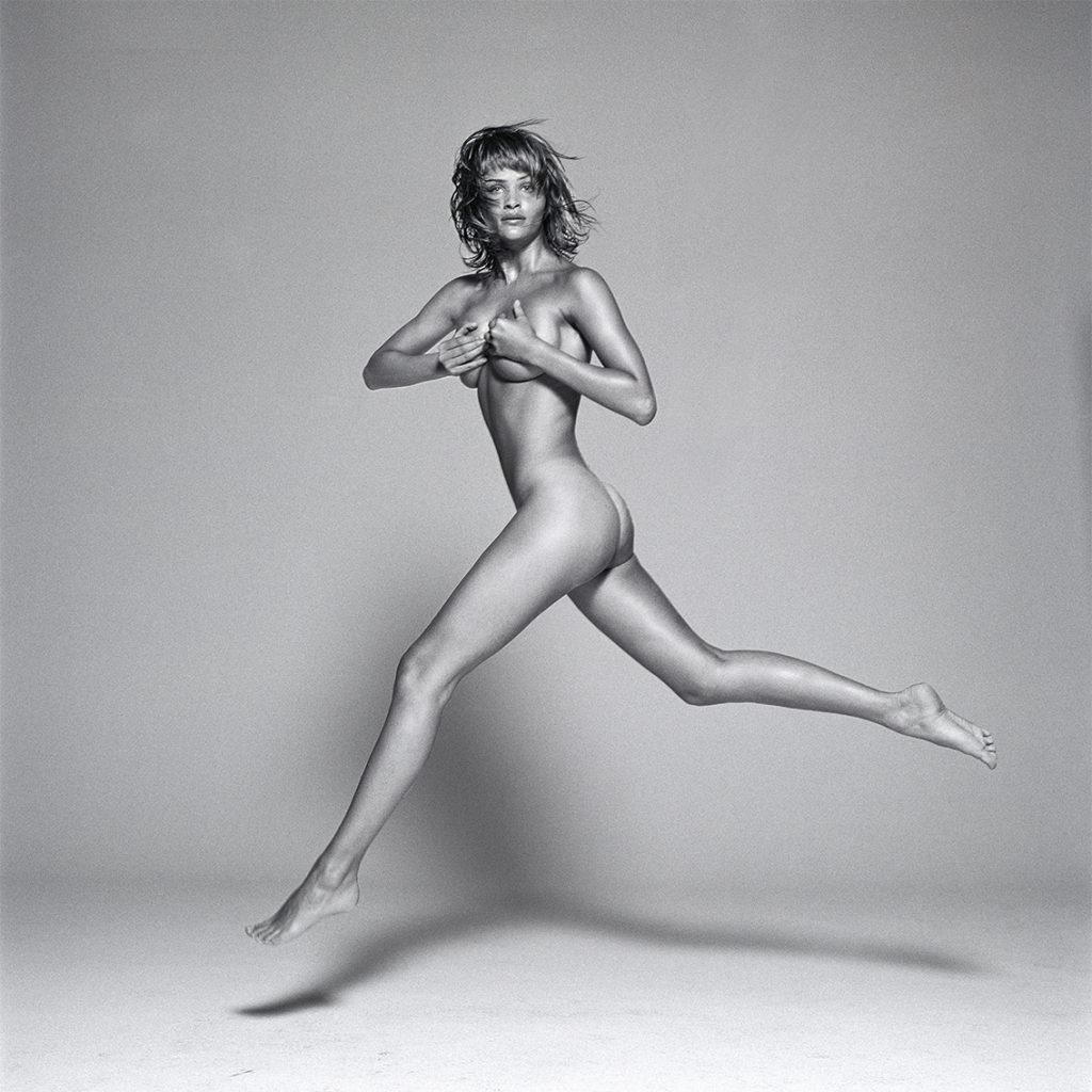 Black and white photo of the nude supermodel covering her breasts with her hands leaping in midair.