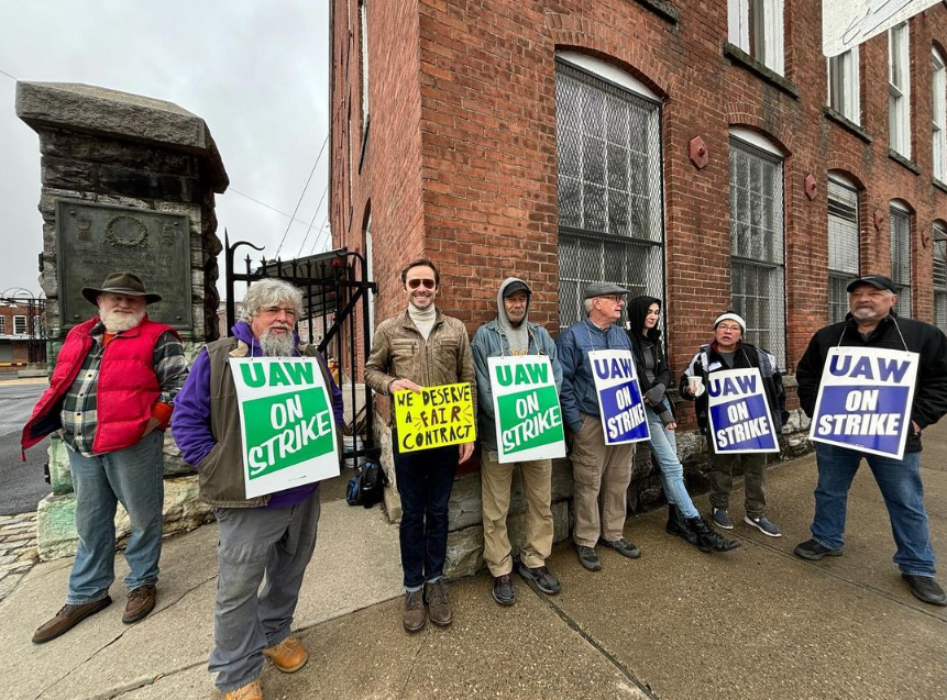 The image shows a group of people, presumably workers, standing outside on a sidewalk near a brick building and a large metal gate, possibly part of an industrial or institutional facility. They are holding protest signs indicating a labor strike. Several signs say "UAW ON STRIKE" in bold, capitalized letters on a white background with a blue border, representing the United Automobile Workers, a labor union. One sign, which stands out because of its yellow background and hand-written style, says "WE DESERVE A FAIR CONTRACT." The individuals are dressed for cool weather. From left to right: the first person is wearing a hat, a red backpack, a purple jacket, and jeans; the second person has a white beard, glasses, and is dressed in a purple jacket and gray pants; the third person, who is holding the yellow sign, is wearing a light brown jacket, dark pants, and glasses; the fourth person is in a gray hoodie and dark pants; the fifth is in a blue jacket and cap; the person next to them is partially obscured but seems to be wearing a dark jacket; and the last person on the right is wearing a black cap, dark jacket, and jeans. The setting looks overcast and the presence of the group suggests they are engaged in a demonstration or picket line related to a labor dispute.