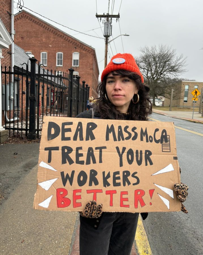 In the image, there is a person holding a protest sign which reads "DEAR MASS MoCA, TREAT YOUR WORKERS BETTER!" The person is standing outdoors, likely on a street sidewalk, with a street and buildings in the background. They are wearing a red beanie hat with a logo, a black coat, and have curly hair. They also appear to be wearing earrings and leopard print gloves. The sign is made of cardboard and the message is written in bold, capital letters. It suggests that the person is advocating for improved treatment of workers at the Massachusetts Museum of Contemporary Art (MASS MoCA).