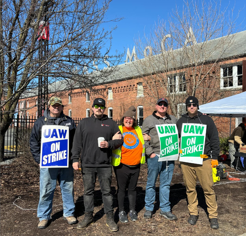 In the image, there is a group of five individuals gathered outdoors during what appears to be a sunny day. They seem to be participating in a strike, as evidenced by the signs they are holding, which read 