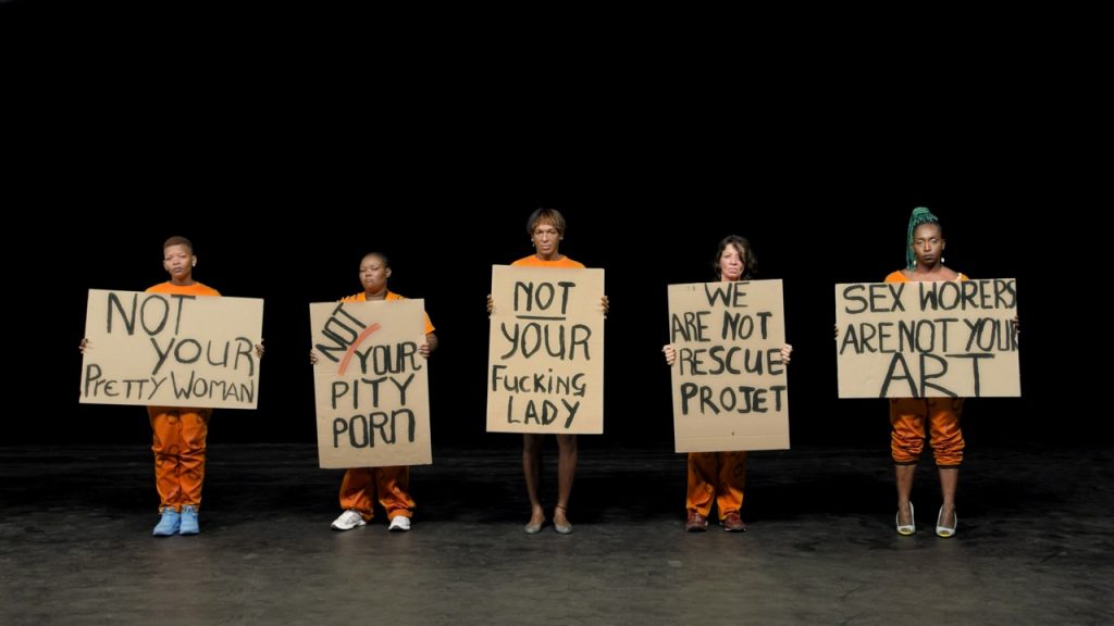 The image shows five individuals dressed in orange outfits standing side by side on a dark background, each holding a large cardboard sign with a handwritten message. From left to right, the signs read "NOT YOUR PRETTY WOMAN," "NOT YOUR PITY PORN," "NOT YOUR FUCKING LADY," "WE ARE NOT YOUR RESCUE PROJECT," and "SEX WORKERS ARE NOT YOUR ART."