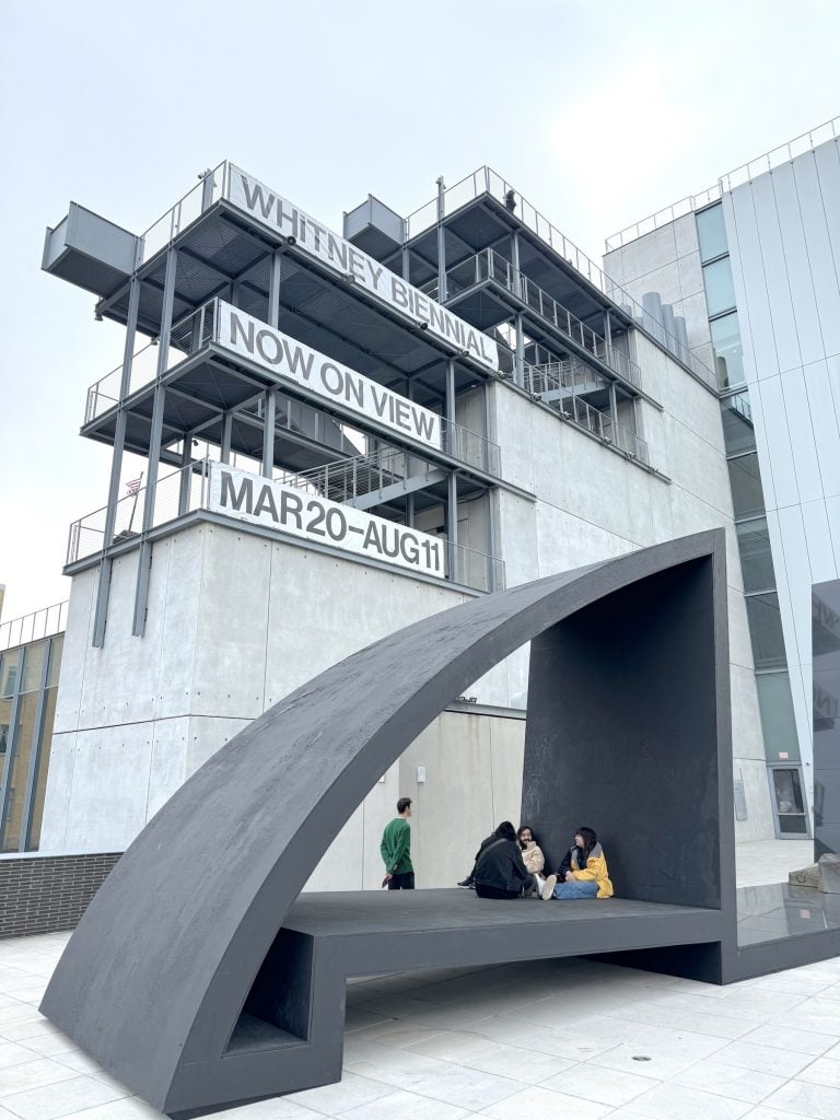 A museum towers above an outdoor terrace featuring a large triangular black sculpture with people sitting on it