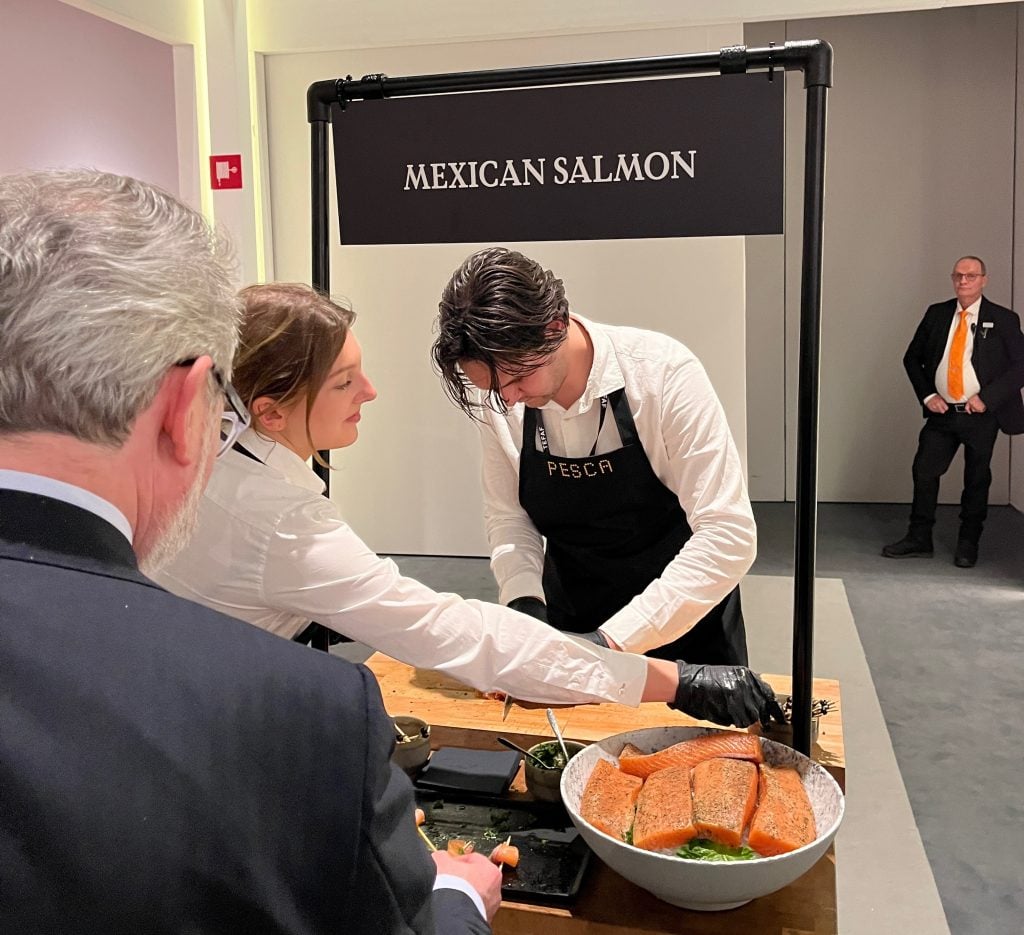 In a color photo, two white-shirt-clad workers prepare orange-pink salmon filets beneath a sign that reads "Mexican salmon"