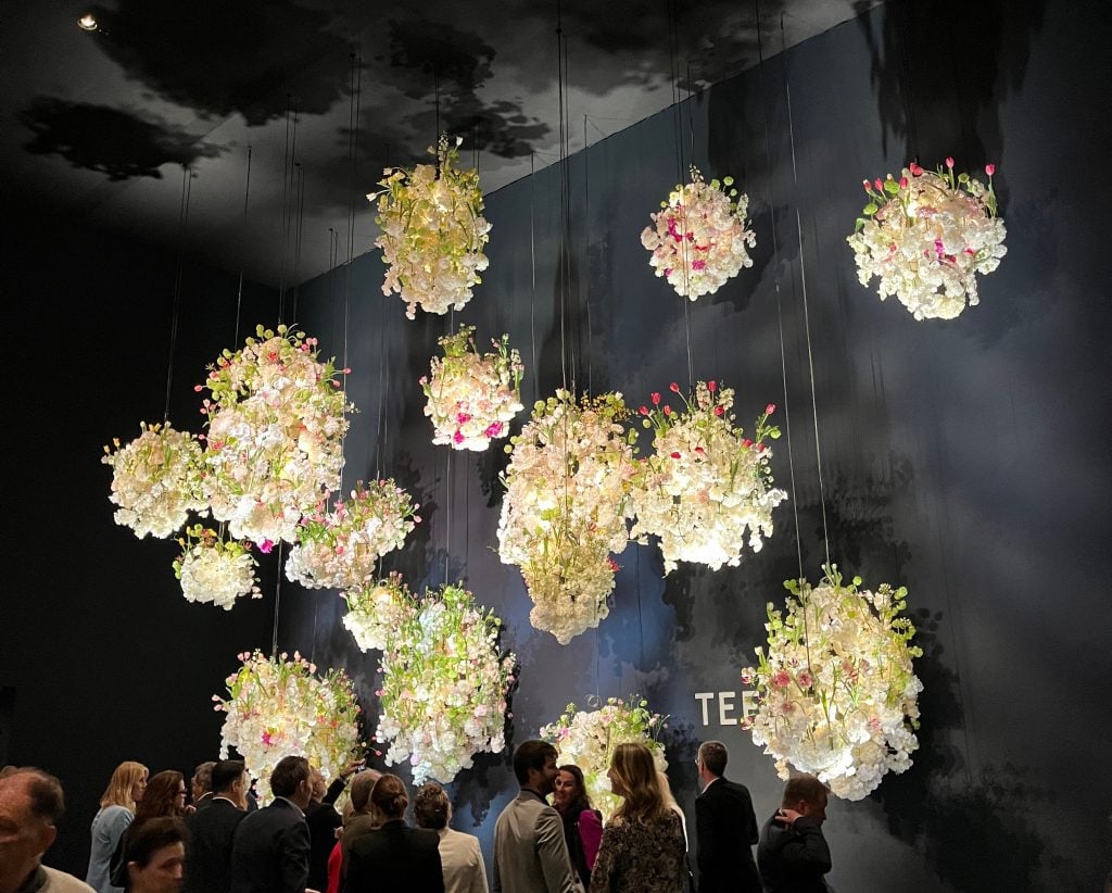 In a color photo, large bouquets of flowers hang from the ceiling in a black-walled space.