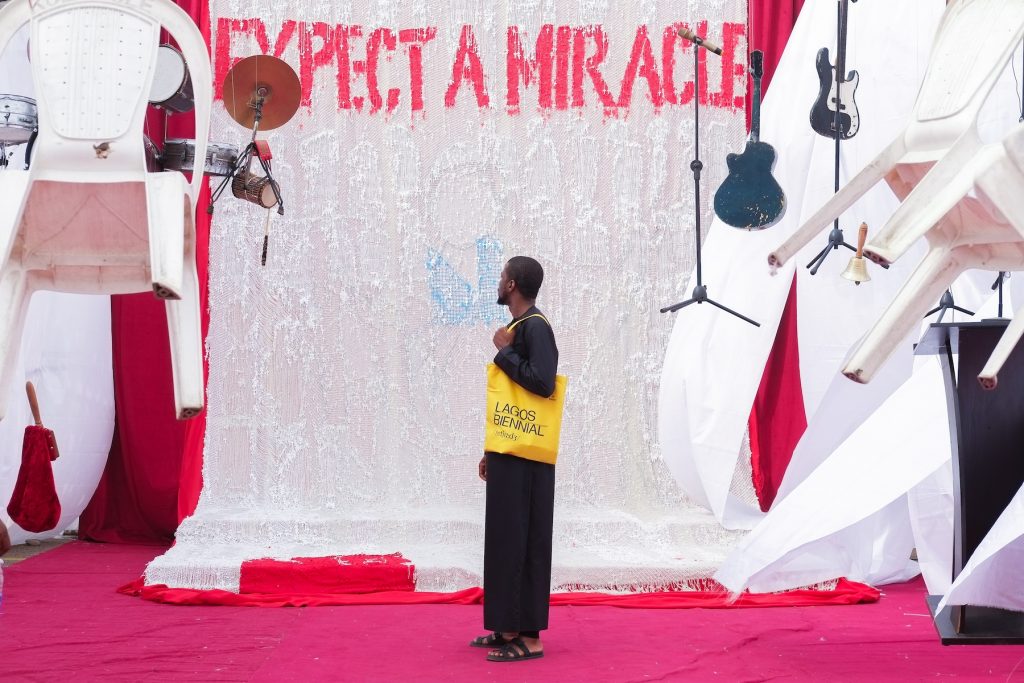 A man with a yellow bag that says LAGOS BIENNIAL stands in front of a wall that says EXPECT A MIRACLEE