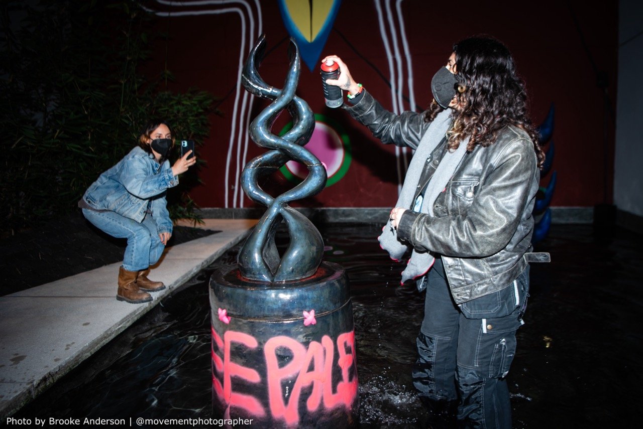 A person interacting with a sculptural fountain with a piece labeled "FREE". They are using a spray paint can on the sculpture, which is set against a glossy black base, reflecting the light and surroundings, as another person captures the moment on their smartphone.